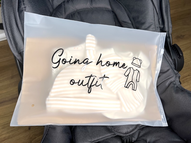 May include: A clear plastic bag with the words "Going home outfit" in black script. Inside the bag are baby clothes, including a striped hat and onesie. A stroller is in the background.