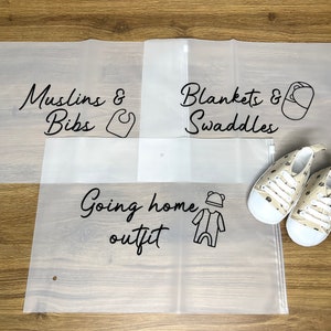May include: Three translucent storage bags labeled "Muslins & Bibs", "Blankets & Swaddles", and "Going home outfit" with corresponding illustrations. A pair of beige baby shoes with a bear pattern is also present.