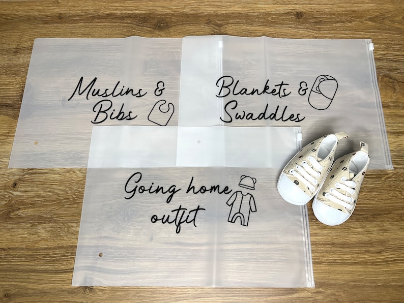May include: Three translucent storage bags labeled "Muslins & Bibs", "Blankets & Swaddles", and "Going home outfit" with corresponding illustrations. A pair of beige baby shoes with a bear pattern is also present.