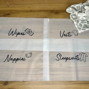 May include: Clear plastic storage bags with black text labels for baby essentials. The labels read "Wipes," "Vests," "Nappies," and "Sleepsuits," each accompanied by a related icon. A gray and white giraffe-patterned blanket and stuffed animal are in the background.