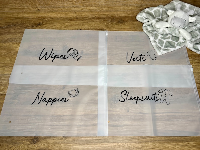 May include: Clear plastic storage bags with black text labels for baby essentials. The labels read "Wipes," "Vests," "Nappies," and "Sleepsuits," each accompanied by a related icon. A gray and white giraffe-patterned blanket and stuffed animal are in the background.