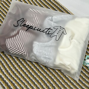 May include: A clear plastic bag labeled "Sleepsuits" contains several baby sleepsuits. The bag is sealed and rests on a striped blanket in shades of gray, white, and yellow. Two blue and white pacifiers are next to the bag.