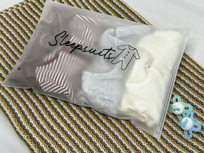 May include: A clear plastic bag labeled "Sleepsuits" contains several baby sleepsuits. The bag is sealed and rests on a striped blanket in shades of gray, white, and yellow. Two blue and white pacifiers are next to the bag.