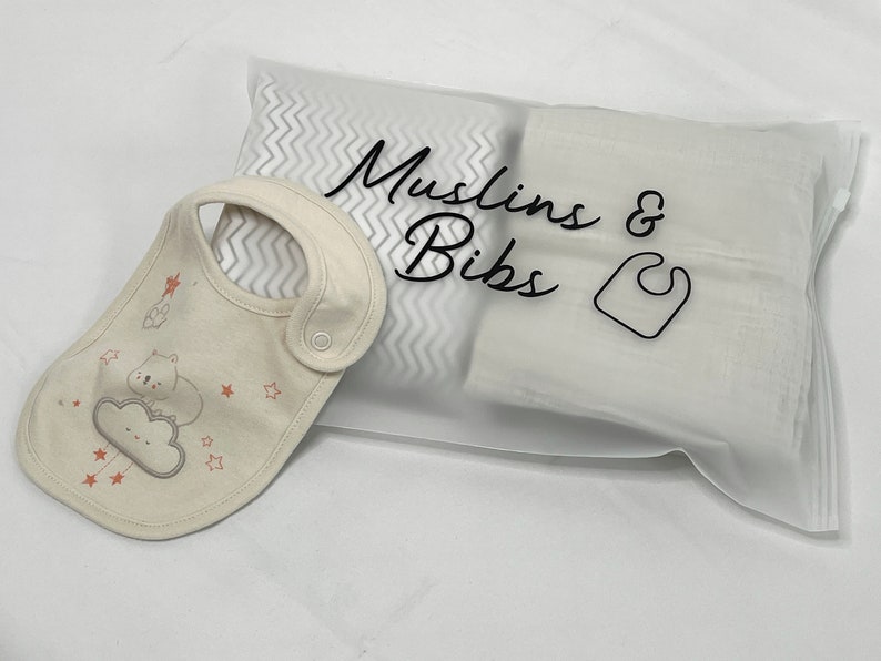 May include: A set of baby essentials, including a bib and muslin cloths, packaged in a clear bag. The bib is beige with a cute cloud and bear design. The bag is labeled "Muslins & Bibs".