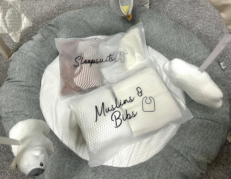 May include: Two clear plastic bags labeled "Sleepsuits" and "Muslins & Bibs" rest on a gray and white play mat. The sleepsuit bag contains striped fabric, while the muslin bag holds white items. A baby bib icon is visible.
