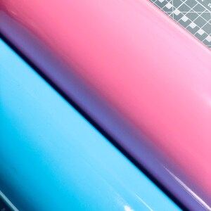 May include: Three rolls of vinyl in shades of blue, pink, and purple. The rolls are positioned diagonally, with a cutting mat and ruler visible in the background. The vinyl has a glossy finish.