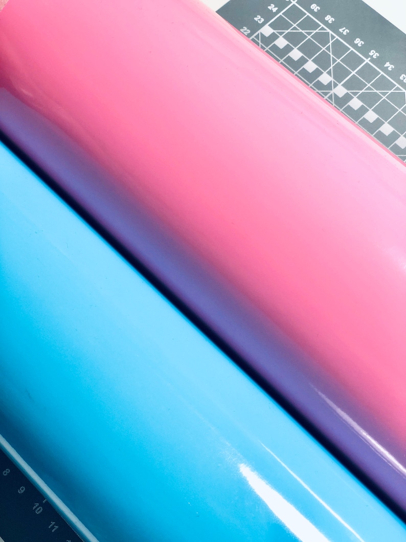 May include: Three rolls of vinyl in shades of blue, pink, and purple. The rolls are positioned diagonally, with a cutting mat and ruler visible in the background. The vinyl has a glossy finish.
