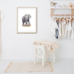 May include: A framed watercolor painting of a gray elephant standing in a field of brown grass. The painting is hanging on a white wall in a room with a white wooden stool and a white shelf with clothes hanging on it.