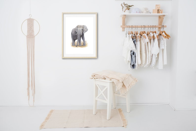May include: A framed watercolor painting of a gray elephant standing in a field of brown grass. The painting is hanging on a white wall in a room with a white wooden stool and a white shelf with clothes hanging on it.