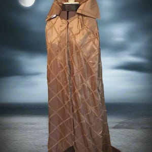 May include: A brown quilted cape with a large collar, displayed on a mannequin. The cape has a diamond pattern and a flowing design. The background features a moon and a seascape, creating a dramatic effect.