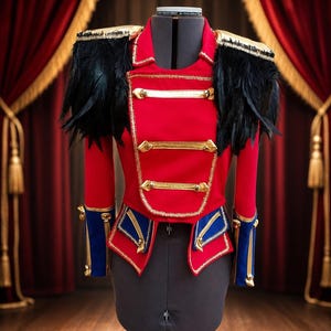 Op de afbeelding: Een rood jasje in militaire stijl met gouden vlechtwerk, zwarte veren epauletten en blauwe manchetten. Het kledingstuk is voorzien van drie horizontale gouden koordsluitingen op de borst en decoratieve gouden accenten op de mouwen.