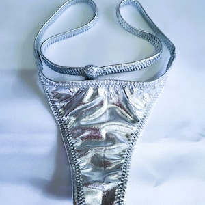 May include: Silver metallic thong-style underwear with thin straps and a high-cut design. The fabric has a crinkled texture and is stitched with a contrasting thread. The underwear is displayed on a white surface.