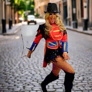 May include: A person in a red, blue, and black costume with a black hat and boots, holding a whip, poses on a cobblestone street. The costume has gold accents and a sequined skirt. The background shows a European street scene.