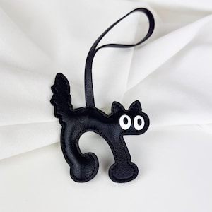 Puede incluir: Llavero de gato de cuero negro con ojos blancos. El gato está en una pose juguetona con la cola enroscada.