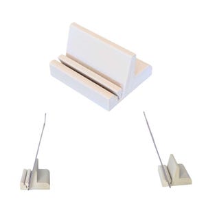 Könnte beinhalten: Ein hellbeige, L-förmiger Telefonständer mit zwei parallelen Rillen zur Aufnahme eines Telefons. Der Ständer besteht aus einem soliden, glatten Material. Zwei zusätzliche Ständer werden mit einer Nadel in der Nut gezeigt.