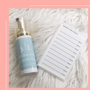 Op de afbeelding: Een witte fles met een blauw etiket waarop "$ | L" en "Wile Lash shampoo by Sparkle Lash" staat, met een gouden dop. De fles staat op een wit, pluche oppervlak naast een wit wimpertray met de tekst "Sparkle Lashes Miami" en metingen in millimeters van 8 mm tot 17 mm.