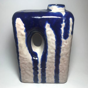 May include: A white ceramic vase with a blue glaze. The vase has a rectangular shape with a hole in the side. The glaze is uneven, creating a textured look.