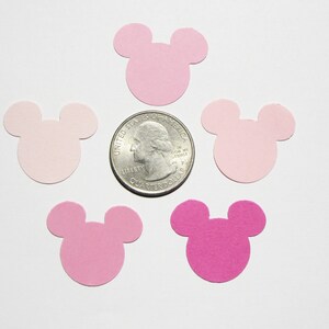 May include: Pink and light pink paper confetti cut in the shape of Mickey Mouse heads. The confetti is scattered on a white surface with a US quarter for scale.