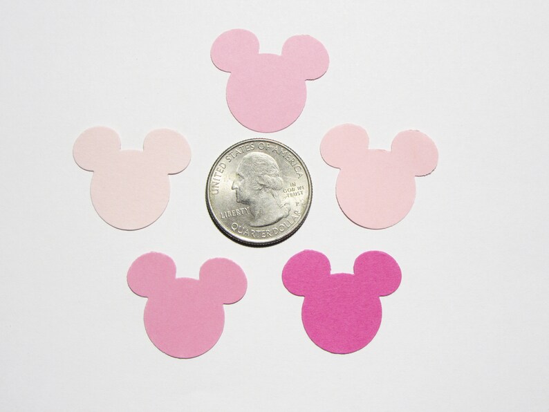 May include: Pink and light pink paper confetti cut in the shape of Mickey Mouse heads. The confetti is scattered on a white surface with a US quarter for scale.