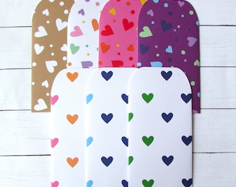 May include: A collection of colorful, heart-patterned cards. The cards feature a variety of colors, including brown, white, pink, red, orange, purple, green, and blue. The hearts are evenly spaced across the surface of each card. The cards are arranged on a white wooden surface.