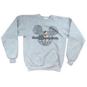 Peut inclure: Sweat-shirt gris avec une silhouette de Mickey Mouse et le texte "Walt Disney World" imprimé sur le devant. La silhouette est composée d'un cercle avec les mots "Magic, Dreams, Tree of Life, Imagination, Adventure, Kingdom, Fantasy, Fun, Memories, Friendship, Family, Together, Since 1971" imprimés autour.