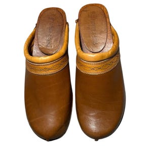 Può includere: Un paio di zoccoli in pelle marrone con finiture marrone chiaro e un motivo decorativo cucito. Gli zoccoli hanno una suola in legno e il testo "THE FATHER & SONS" è visibile sulla soletta.