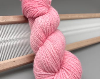 May include: A skein of soft, light pink yarn, tightly wound, rests on a wooden frame with white vertical slats. The yarn is a solid color, with a slightly textured appearance. The background is a solid gray.