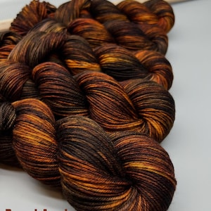 May include: A collection of hand-dyed yarn skeins featuring a variegated color palette of deep espresso brown, burnt orange, and golden yellow tones. The text "Barrel-Aged" is printed in brown letters at the bottom left of the image.