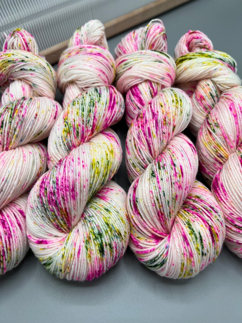 May include: Four skeins of yarn in a white base with pink, green, and yellow speckles. The yarn is arranged in a row, with each skein twisted into a loop.