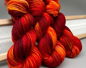 May include: Three skeins of hand-dyed yarn featuring a vibrant variegated color palette of deep crimson, bright orange, and golden yellow. The yarn is twisted into a traditional hank shape, showcasing a smooth, plied texture throughout the fibers.