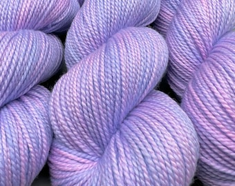 May include: Three skeins of soft, purple yarn with a subtle pink ombre effect. The yarn is arranged in a loose, overlapping pattern.
