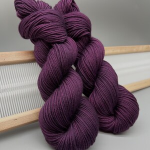 May include: Two skeins of deep purple yarn are shown, highlighting their twisted structure. The yarn is a rich, solid colour, and looks soft and tightly spun. The skeins are resting on a wooden weaving tool, with a white background.