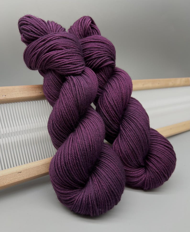May include: Two skeins of deep purple yarn are shown, highlighting their twisted structure. The yarn is a rich, solid colour, and looks soft and tightly spun. The skeins are resting on a wooden weaving tool, with a white background.