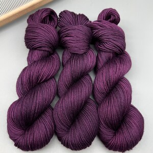 May include: Three skeins of deep purple yarn are shown, each tightly wound into a twisted shape. The yarn has a smooth, lustrous appearance, suggesting a high-quality fibre. A wooden weaving tool is visible in the background.