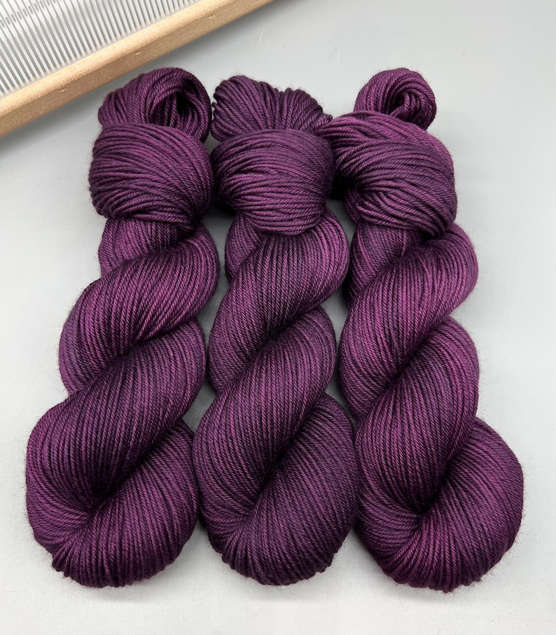 May include: Three skeins of deep purple yarn are shown, each tightly wound into a twisted shape. The yarn has a smooth, lustrous appearance, suggesting a high-quality fibre. A wooden weaving tool is visible in the background.