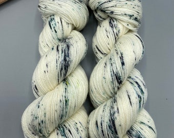May include: Two skeins of white yarn with black and green speckles. The yarn is arranged in a twisted pattern.