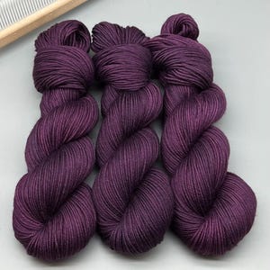 May include: Three skeins of deep purple yarn are shown, each tightly wound into a compact bundle. The yarn has a smooth, lustrous appearance, suggesting a high-quality fibre. A wooden knitting tool is visible in the upper left corner, hinting at the yarn's use in crafting.