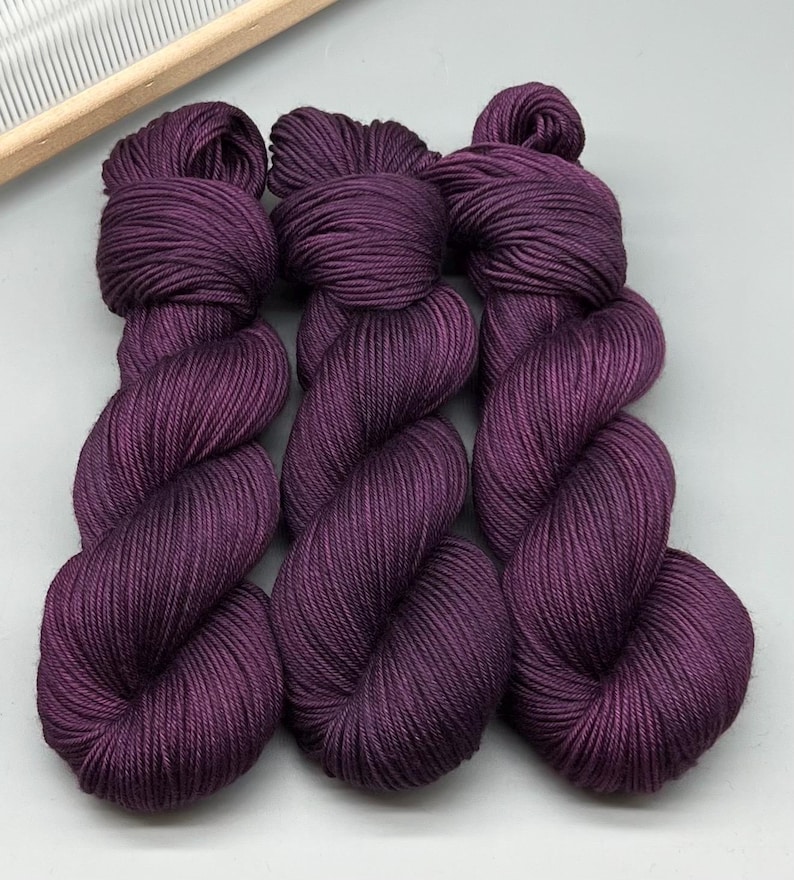 May include: Three skeins of deep purple yarn are shown, each tightly wound into a compact bundle. The yarn has a smooth, lustrous appearance, suggesting a high-quality fibre. A wooden knitting tool is visible in the upper left corner, hinting at the yarn's use in crafting.
