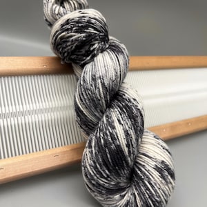 May include: A skein of speckled yarn in black, white, and gray, resting on a wooden weaving frame. The yarn is tightly twisted, showcasing a blend of colors. The wooden frame has a series of vertical white threads, suggesting a craft project.