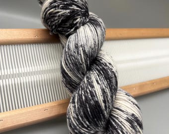 May include: A skein of speckled yarn in black, white, and gray, resting on a wooden weaving frame. The yarn is tightly twisted, showcasing a blend of colors. The wooden frame has a series of vertical white threads, suggesting a craft project.
