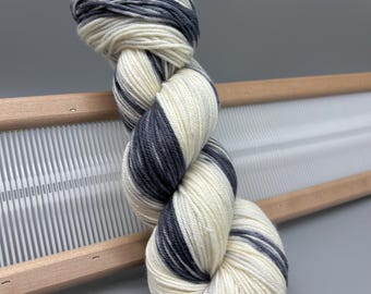 May include: A skein of white and charcoal grey yarn with a striped pattern. The yarn is wound into a ball and is sitting on a wooden loom.