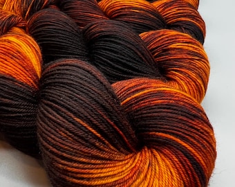 May include: A skein of yarn featuring a variegated color pattern of deep espresso brown and vibrant sunset orange. The yarn is tightly twisted into a thick, multi-layered coil, showcasing a smooth, plied texture throughout the entire length.