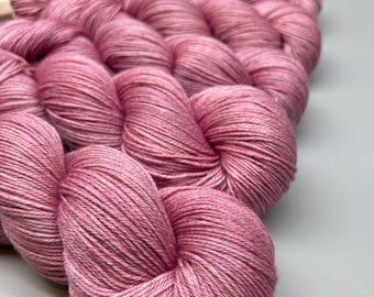 May include: Close-up of pink yarn skeins arranged on a weaving loom. The yarn is a soft, dusty rose color, with visible strands. The texture of the yarn is smooth and tightly wound. The loom is made of wood and has a white grid.