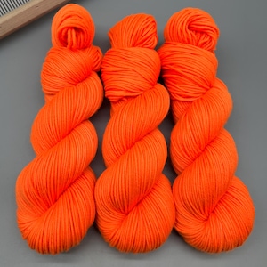 May include: Three skeins of bright orange yarn are displayed against a gray background. The yarn is tightly wound into a twisted shape, with a rounded top and a long, flowing body. The vibrant color and texture suggest a craft or textile project.