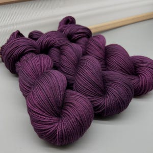 May include: Close-up of several skeins of deep purple yarn. The yarn is tightly wound and has a smooth texture. A wooden frame is visible in the background, suggesting a crafting or knitting project.