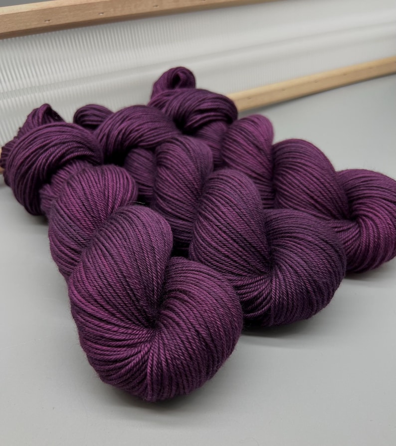 May include: Close-up of several skeins of deep purple yarn. The yarn is tightly wound and has a smooth texture. A wooden frame is visible in the background, suggesting a crafting or knitting project.