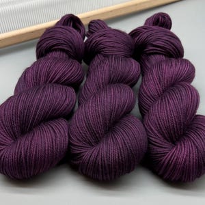 May include: Nine skeins of deep purple yarn are arranged together. The yarn is tightly wound, with a smooth texture. A wooden knitting needle holder is visible in the background, suggesting a crafting theme.