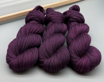 May include: Nine skeins of deep purple yarn are arranged in a cluster. The yarn is tightly wound, showing a smooth texture. A wooden knitting needle holder is visible in the background, suggesting a crafting theme.