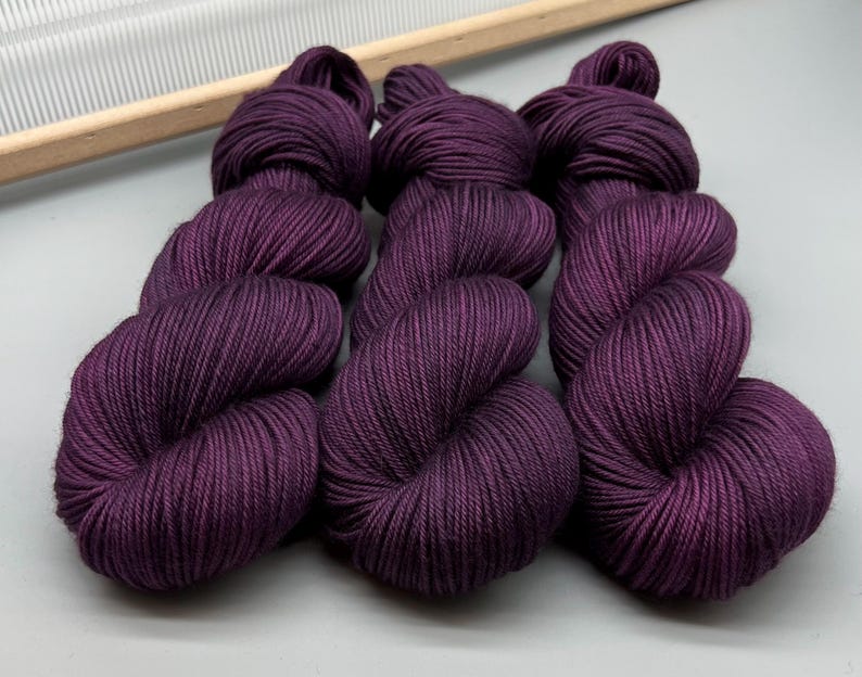 May include: Nine skeins of deep purple yarn are arranged together. The yarn is tightly wound, with a smooth texture. A wooden knitting needle holder is visible in the background, suggesting a crafting theme.