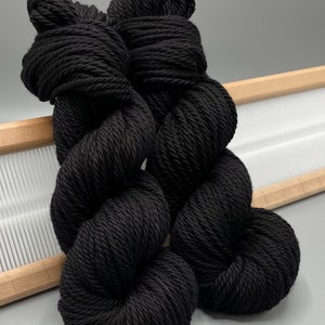 May include: Two skeins of black yarn, likely wool, are shown in a close-up. The yarn is twisted and piled on top of a wooden loom.